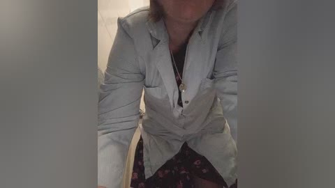 Video of a woman with light skin, wearing a partially unbuttoned light gray jacket over a black floral dress. She stands in a dimly lit hallway, with a blurred background.