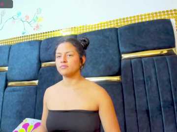 Video of a woman with dark hair in a bun, wearing a strapless black dress, standing in front of a blue upholstered headboard with gold accents and a colorful, floral-patterned wall.