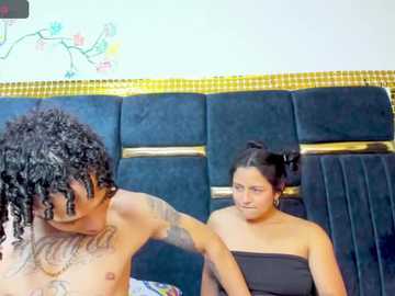 Video of a shirtless, tattooed man with dreadlocks adjusting the strap of a strapless black dress on a woman with dark hair in a bun, sitting on a plush, blue velvet couch with gold trim.