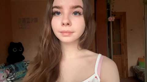 Video of a young woman with long brown hair, blue eyes, wearing a white spaghetti strap top, standing in a cozy, softly lit bedroom with a plush black cat toy, a colorful bedspread, and a wooden door in the background.