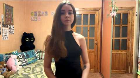 Video of a young woman with long brown hair, fair skin, and a slim physique, wearing a black halter dress, standing in a cozy, cluttered bedroom with wooden doors and a green patterned couch.