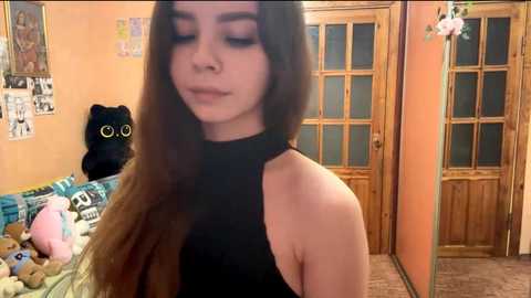 Video of a young woman with long brown hair, wearing a black halter top, standing in a cozy room with a wooden door, a stuffed animal, and a cat poster.