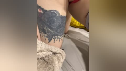 Video of a person's upper thigh and lower hip, showcasing a detailed black lace tattoo with intricate floral designs and dangling chains. The individual is wearing a red bra and laying on a beige bed with a yellow pillow.