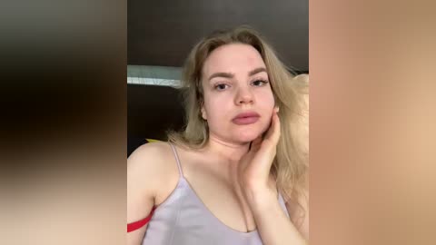 Video of a fair-skinned, blonde woman with wavy hair, wearing a light purple spaghetti-strap top, resting her hand on her cheek. Background includes a blurred, dark-colored wall and a partially visible yellow object.
