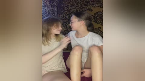 A video captures two young women with light skin and shoulder-length hair, sitting on a red carpet. They are dressed casually, with one wearing glasses. They are smiling and touching each other's hands. The background features a patterned tapestry.