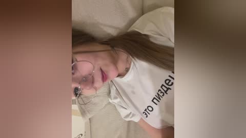 A close-up video of a young woman with long brown hair, wearing glasses and a white t-shirt, lying on a beige couch. Her facial expression is neutral.