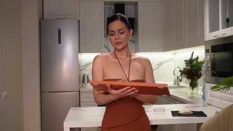 Video of a woman in a strapless, orange dress, holding a wooden box, in a modern, light-colored kitchen with stainless steel appliances and a marble backsplash.