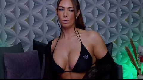 Video of a voluptuous woman with large breasts, tan skin, and long brown hair, wearing a black bikini top and black robe, sitting on a grey couch against a floral-patterned wall.