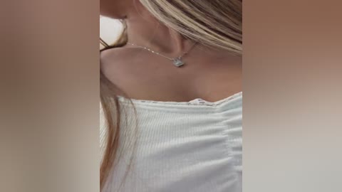 A close-up video of a young woman with long, straight blonde hair, wearing a white ribbed tank top and a delicate silver necklace with a small pendant. The image is soft-focused, with a warm, blurred background.