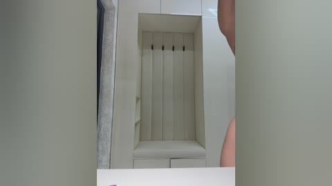 Video of a minimalist, modern closet with white walls and a cushioned bench. A gray coat hangs on the left, and a person's hand is visible on the right. The interior features vertical bars and a small drawer.