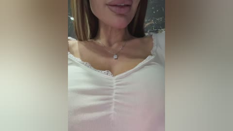 Video of a light-skinned woman with long, straight brown hair, wearing a white, ruffled, off-the-shoulder top with a lacy neckline. She has a delicate necklace with a small diamond pendant. Background is blurry, featuring a patterned cushion.