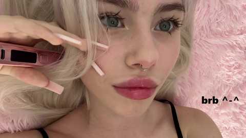 Video of a young woman with platinum blonde hair, blue eyes, and full lips. She's wearing a nose ring, a black bra, and is holding a pink phone. Pink fluffy background.