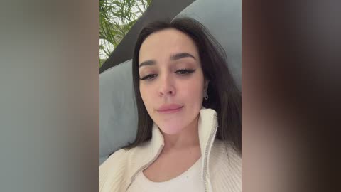 Video of a young woman with long, straight dark hair, fair skin, and light makeup, wearing a white ribbed jacket, standing against a grey wall with green foliage in the background.