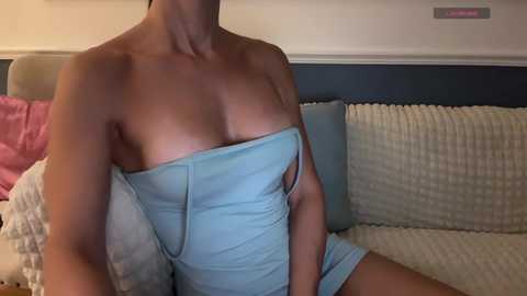 Video of a light-skinned, shirtless woman with large breasts, wearing a blue strapless dress, sitting on a beige sofa with white and pink pillows, in a dimly lit room.