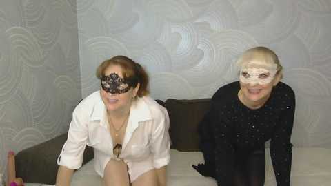 Video of two women in white shirts, black lace masks, and black gloves, kneeling on a bed with floral wallpaper in the background.