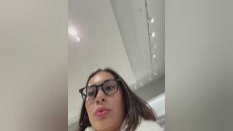 A video of a young woman with straight brown hair, wearing black-rimmed glasses, and a white shirt, taken from an upward angle in a white, modern interior.