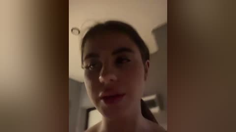 A video of a young woman with fair skin, brown eyes, and dark hair, smiling slightly. She's indoors, with blurred background elements.