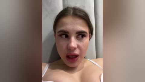 A video of a young woman with light skin, dark hair, and a surprised expression. She wears a white tank top, her breasts are visible. The background features a beige curtain.