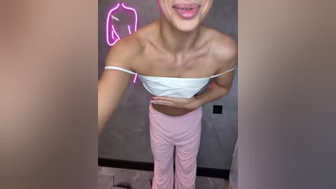 Video of a slim, light-skinned woman with short blonde hair, wearing a white off-shoulder crop top and light pink pants, smiling. Neon horse silhouette on gray wall.