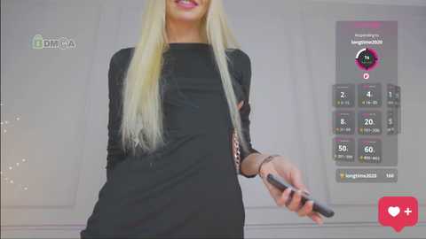 Video of a blonde woman in a black dress holding a smartphone, standing against a white background. The image includes a virtual \"like\" counter and social media icons.