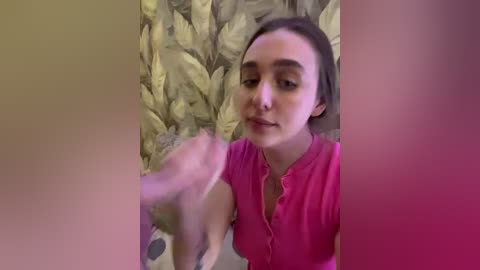 Video of a young woman with light skin and straight brown hair, wearing a pink blouse, looking at a blurry object in front of her. Background features green leaf-patterned wallpaper.