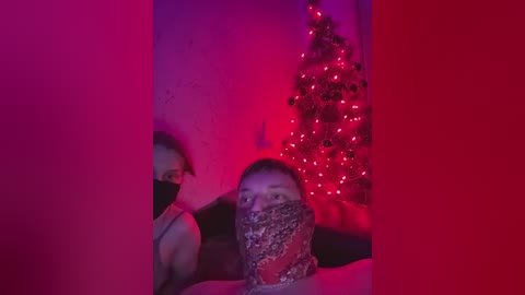 A dimly-lit video of a man with a black mask and a woman with a leopard-print mask, both in black tank tops, in a room with a lit Christmas tree and red lighting.