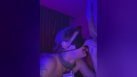 Video of a man with light skin, wearing a black blindfold and collar, kneeling in a dimly lit room with purple and red lighting, tattoo on his shoulder, and holding a black object.