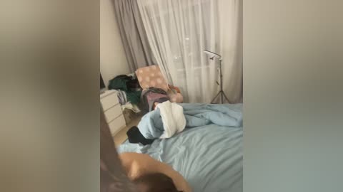 A video of a messy bedroom with a beige bed, a white dresser, a green chair, and a fan; a person, partially obscured, lies on the bed.