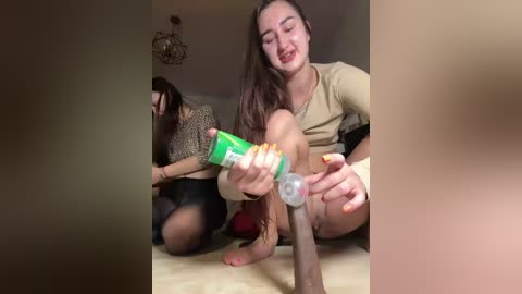 Video of two women in a bedroom: one using a green vibrator on a man's erect penis, while the other sits nearby.