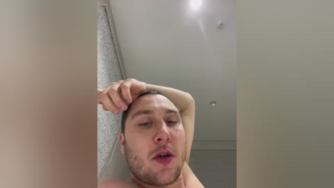 Video of a young, light-skinned man with short hair and a beard, standing shirtless, rubbing his head in a bathroom with a white ceiling and a silver shower curtain.