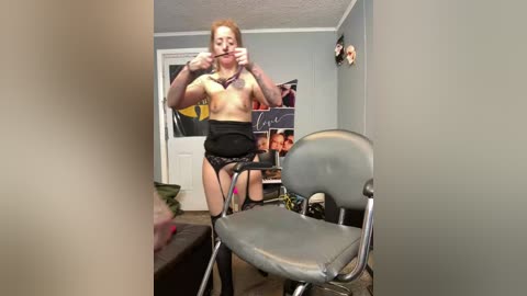 Video of a slender, fair-skinned woman with red hair, wearing black lingerie, holding her mouth open in a bedroom with posters and a chair.