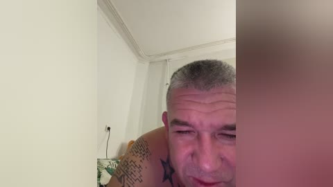 Video of a middle-aged, balding man with a muscular build, visible tattoos, and light skin, leaning against a white wall with a slight smile. Background includes a ceiling with crown molding and a green plant.