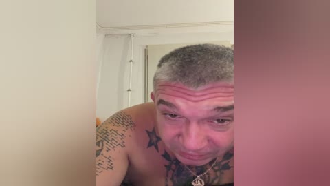 Video of a shirtless man with short gray hair, wearing a necklace, and tattoos including a star pattern, captured in a close-up, low-resolution selfie. Background shows a plain white wall with a partially visible door.