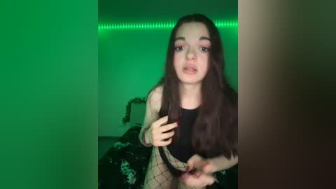 Video of a young woman with long brown hair, fair skin, and green eyes, wearing a black crop top and fishnet stockings, posing indoors with green LED lights in the background.