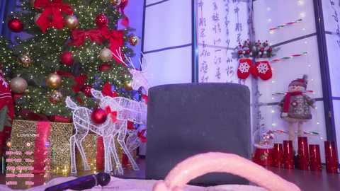 Video of a festive holiday setting: a decorated Christmas tree, red reindeer, white reindeer, and a gray chair.