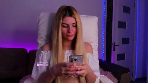 A video of a blonde Caucasian woman in a white off-shoulder dress, seated on a brown couch, smiling at her smartphone, with purple LED lighting in the background.