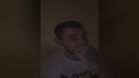 A dimly-lit video of a young man with a short beard, wearing a white t-shirt, standing behind a wooden door, holding his hand to his mouth in a secretive manner.