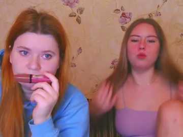 A video shows a young redhead in a blue top eating a red popsicle, while a young woman with long brown hair in a purple top looks on. The background features a floral-patterned wall.