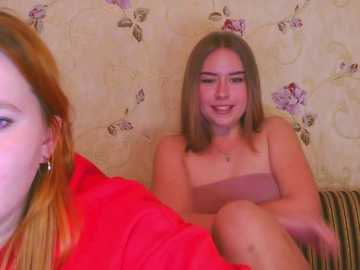 Video of a fair-skinned, slim young woman with long, straight auburn hair and medium-sized breasts, wearing a strapless top, sitting on a striped couch against a floral wallpaper background.