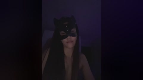 A dimly lit video of a person with long brown hair, wearing a black cat mask and a black top, set against a dark, purple-toned background.