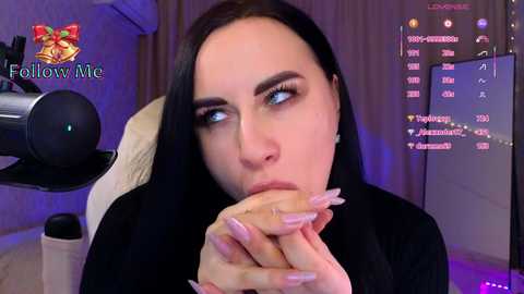 A video shows a fair-skinned woman with long, dark hair, wearing a black outfit, eating a donut, with a gaming setup in the background.