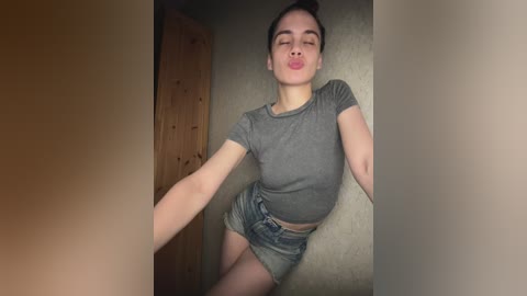 A video of a young woman with light skin, brown hair tied in a bun, wearing a tight gray crop top and denim shorts, sticking her tongue out and smiling. She's in a dimly lit room with wooden and beige walls.