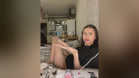 Video of a young woman with light skin, dark hair, and a slender physique, sitting on a bed, holding a phone, in a cluttered, dimly-lit bedroom with a kitchen in the background.