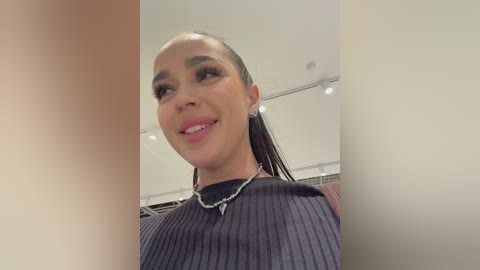 Video of a young woman with medium skin tone, straightened black hair, and dark eyebrows. She wears a black ribbed top with a silver necklace, smiling in an indoor setting with white walls and recessed lighting.