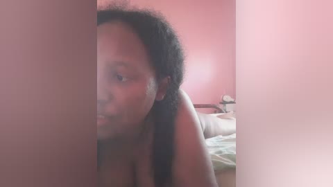 Video of a topless Black woman with curly hair partially obscured by a pink wall, standing in a dimly lit room with a bed in the background.