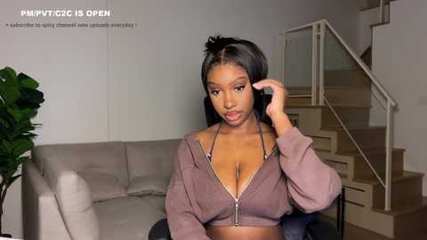 Video of a young Black woman with long black hair, wearing a pink cropped hoodie, sitting on a beige couch in a modern living room.