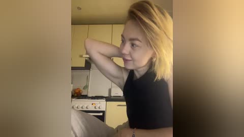 A video of a fair-skinned woman with blonde hair, wearing a black shirt, in a modern kitchen with beige cabinets and a white stove.