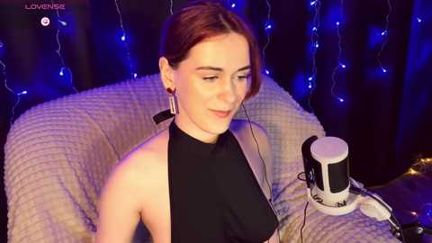 Video of a fair-skinned, red-haired woman in a black halter dress, wearing headphones, sitting on a textured, beige chair with blue fairy lights in the background, in a cozy indoor setting.