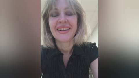 Video of a smiling, middle-aged Caucasian woman with shoulder-length blonde hair, wearing a black, short-sleeved blouse. The background is blurred, with a soft, neutral color palette.