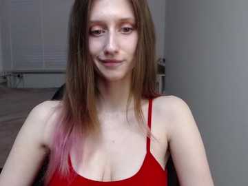 Video of a young woman with long, straight, light brown hair, fair skin, and a slender physique. She wears a red spaghetti-strap top, standing indoors with white blinds and a radiator in the background.